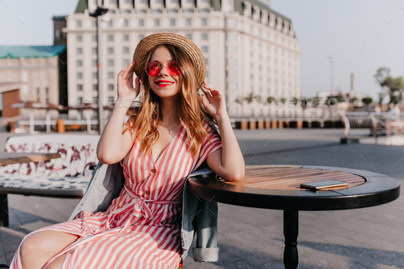 Fascinating white girl in straw hat sitting in outdoor cafe. Portrait of blithesome european lady in beautiful striped dress chilling on city background.